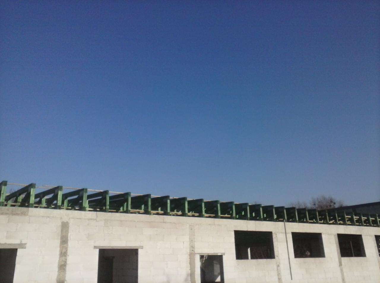 Surowy stan budynku z białych bloczków z zaznaczonymi otworami okiennymi i drzwiowymi oraz konstrukcją dachu z zielonych belek na tle błękitnego nieba.