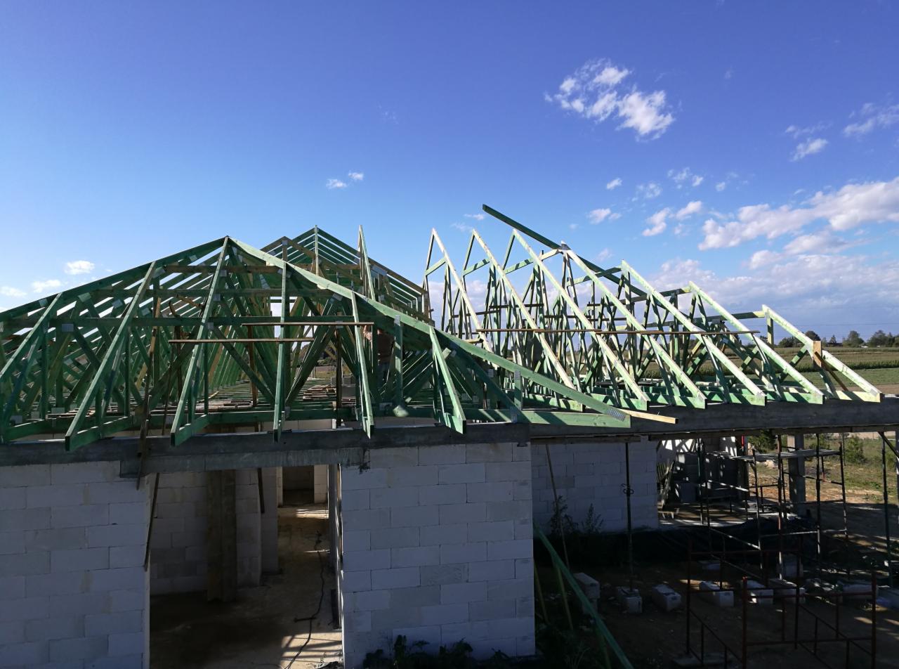 Wznoszenie konstrukcji dachu z impregnowanego drewna na murowanym domu z białych bloczków, widok na niebo z lekkimi chmurami.