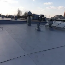 Płaski dach przemysłowy z szarej membrany PVC, widoczne kominki wentylacyjne, świetlik dachowy, instalacja odgromowa i kominy elektrowni w tle.