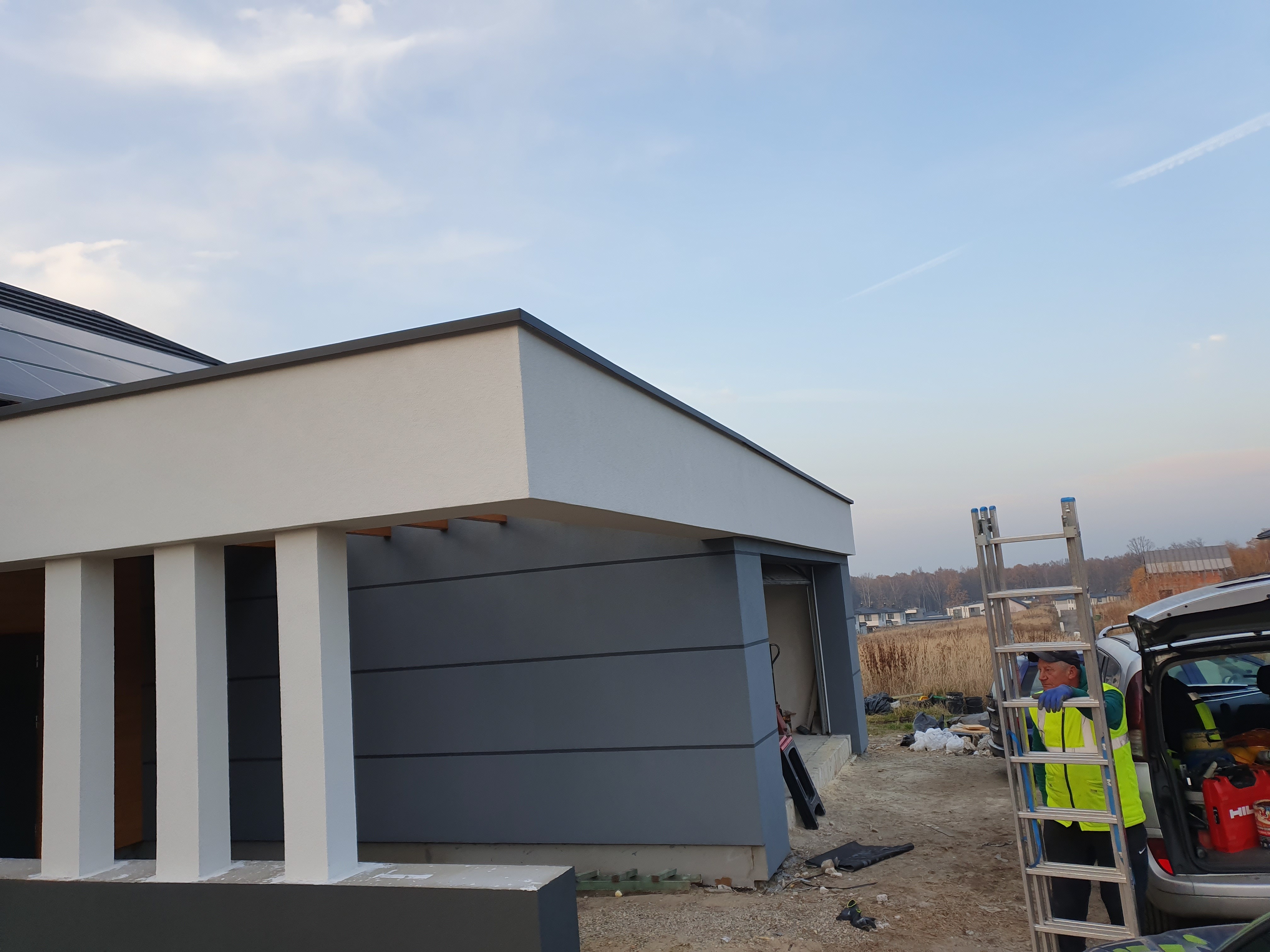 Wykończony budynek z obróbkami blacharskimi, widoczny pracownik z drabiną i otwarty bagażnik samochodu w tle.