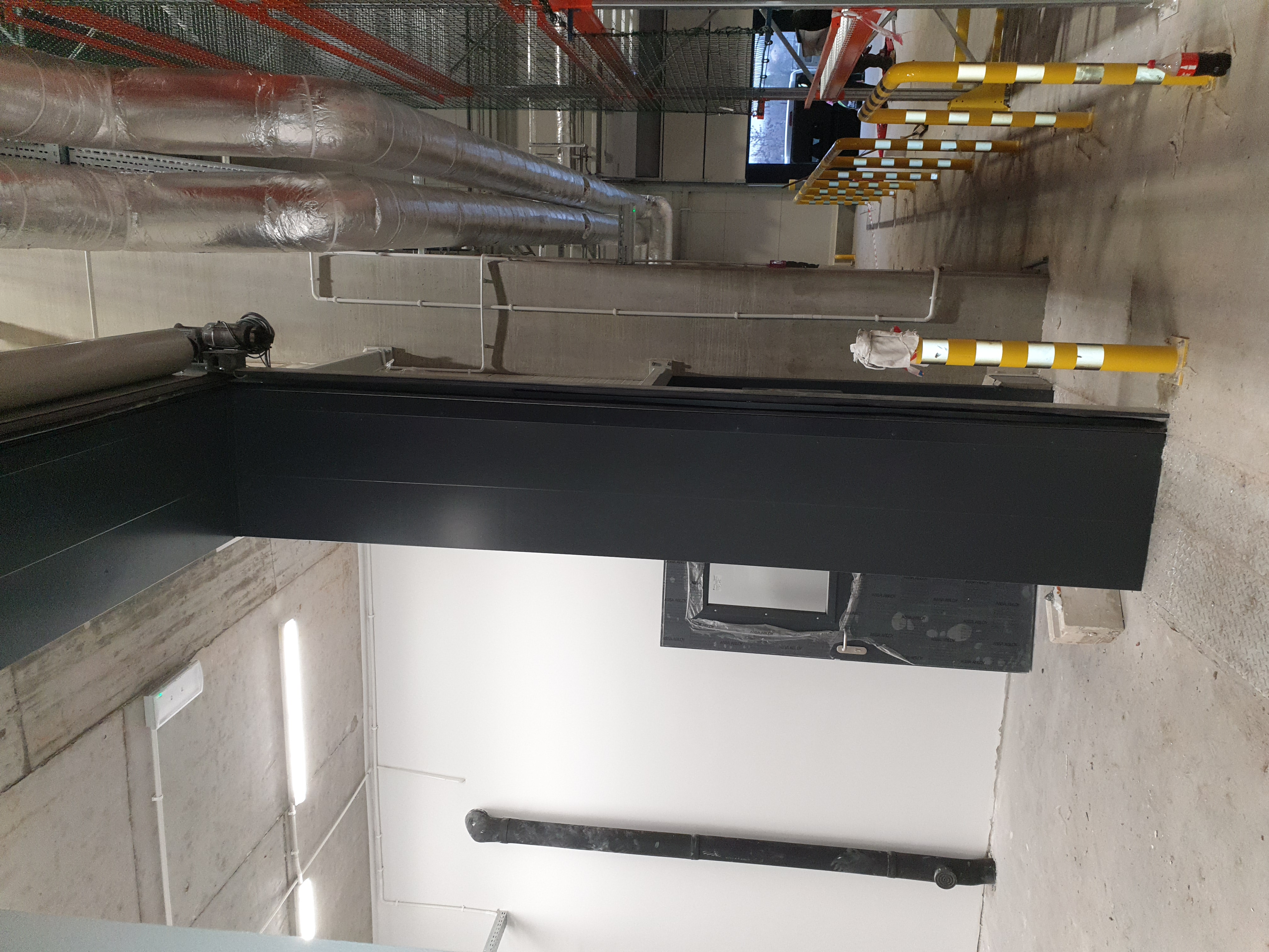 Wnętrze garażu podziemnego z widocznymi rurami wentylacyjnymi pod sufitem, zabezpieczeniami w postaci żółto-białych słupków oraz instalacją elektryczną.