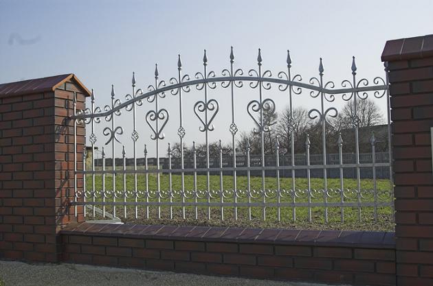 Kuta, metalowa balustrada ogrodzeniowa z motywem serc i zdobieniami, osadzona na niskim murku z cegły klinkierowej w Zielonkach, z widokiem na trawnik i drzewa w tle.