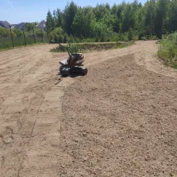 Przygotowanie podłoża pod trawnik: ubijarka gleby na piaskowej ziemi, częściowo wyrównany teren, w tle zieleń i domy.