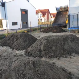 Świeżo przywieziona ziemia usypana w pryzmy na piaszczystym podłożu, w tle nowoczesny dom i wywrotka z podniesioną skrzynią ładunkową oraz trampolina.