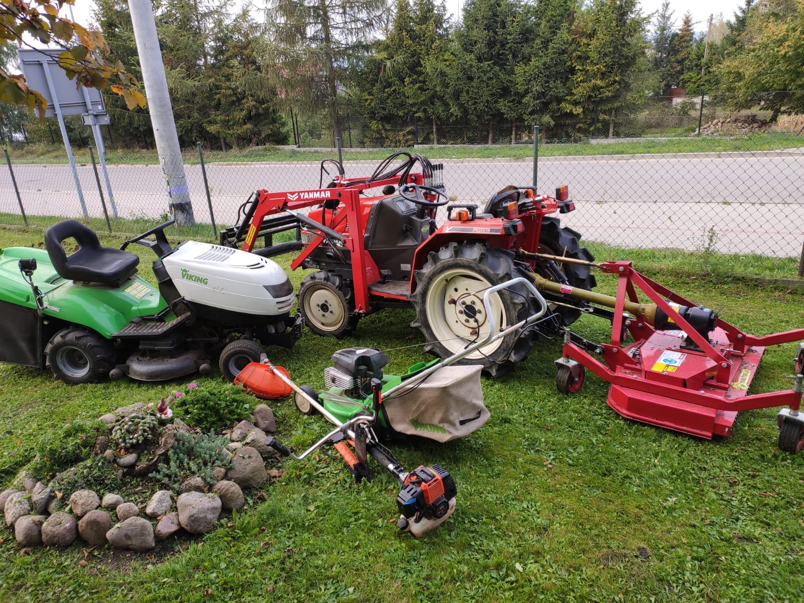 Różnorodny sprzęt ogrodniczy: czerwony traktor Yanmar z kosiarką bijakową, kosiarka samojezdna zielona i biała Viking, podkaszarka spalinowa leżą na trawie obok skalniaka z roślinami.