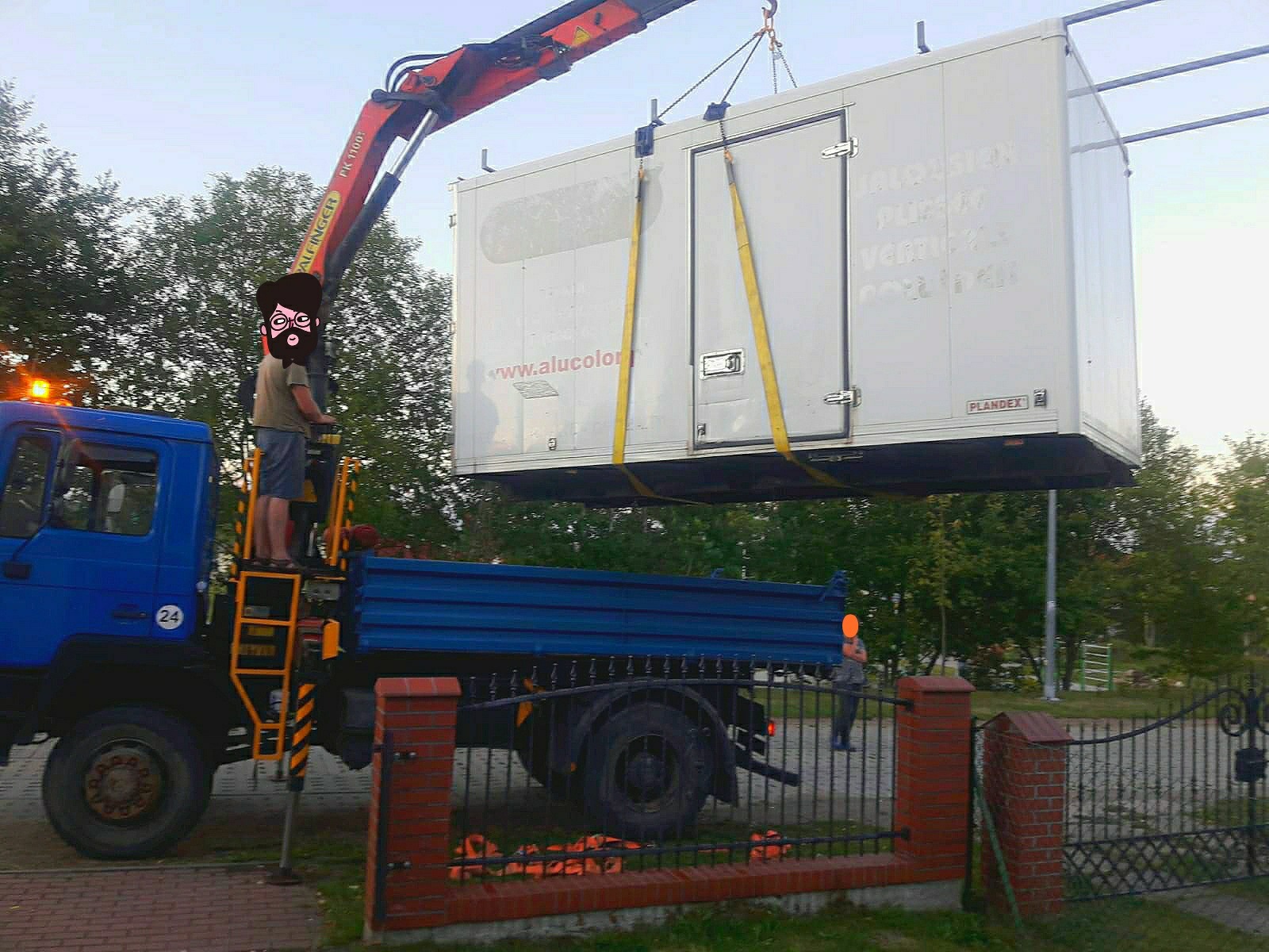 Niebieska ciężarówka z dźwigiem podnosi biały kontener z drzwiami, widoczny operator na platformie i fragment ogrodzenia w tle.