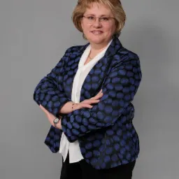 Portret kobiety w okularach, białej bluzce i niebieskiej marynarce w kropki na szarym tle, z założonymi rękami i zegarkiem na nadgarstku.