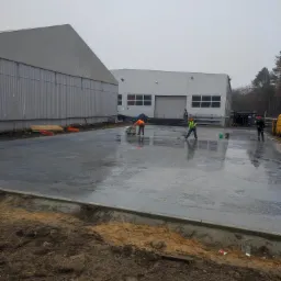 Wylewka betonowa na placu budowy, dwóch pracowników w odblaskowych kamizelkach wygładza powierzchnię za pomocą zacieraczki i rakli, w tle budynki magazynowe i pochmurne niebo.