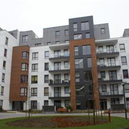 Inwest-Profil Sp.Z O. O. Sp. k. - Nowoczesny budynek wielorodzinny z balkonami, otynkowany na biało z elementami w kolorze szarym i ceglastym, widok z poziomu trawnika z zaparkowanym białym busem.