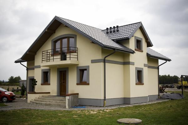 Nowoczesny dom jednorodzinny z beżową elewacją, szarą podmurówką i grafitową dachówką, widoczne brązowe okna i drzwi wejściowe, balkon z czarną balustradą, na trawniku studzienka rewizyjna.
