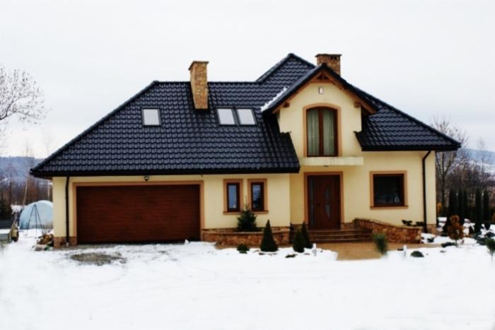 Dwupiętrowy dom z beżową elewacją, czarnym dachem, brązową bramą garażową i oknami, otoczony śniegiem w zimowy dzień.