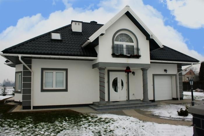 Biały dom z czarnym dachem w zimowej scenerii, widoczne okna z szarymi ramami i garaż z bramą segmentową.