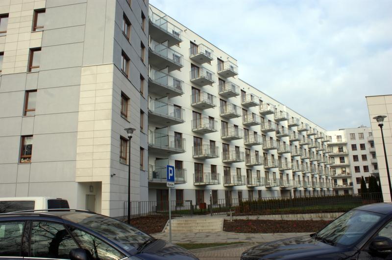 Nowoczesny budynek wielorodzinny z białymi elewacjami i balkonami z przeszklonymi balustradami, widok z poziomu ulicy z zaparkowanymi samochodami.