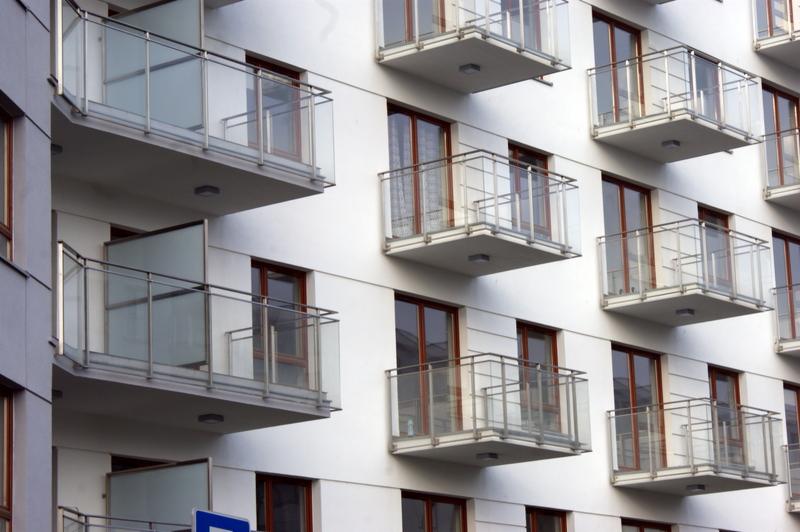 Nowoczesny budynek z białymi ścianami i balkonami z przeszklonymi balustradami, widoczne brązowe ramy okienne.