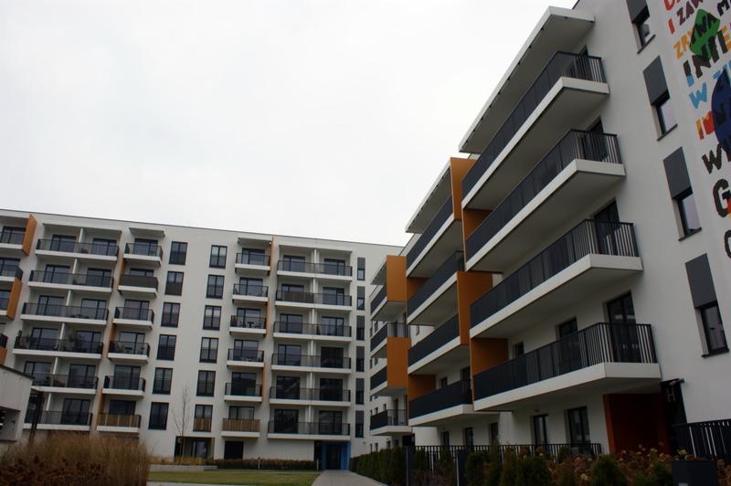 Nowoczesny budynek mieszkalny z balkonami i oknami, widok z dołu na fasadę z elementami dekoracyjnymi i częściowo zasłoniętymi oknami.