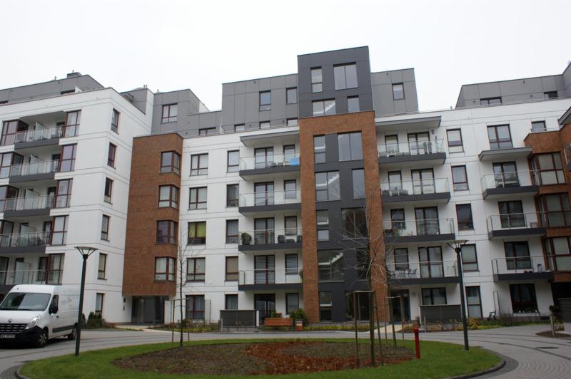 Nowoczesny budynek wielorodzinny z balkonami, otynkowany na biało z elementami w kolorze szarym i ceglastym, widok z poziomu trawnika z zaparkowanym białym busem.