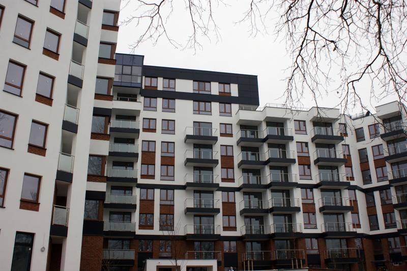 Fasada nowoczesnego budynku wielorodzinnego z oknami PCV i aluminiowymi, balkony z szklanymi balustradami, widok z dołu na tle pochmurnego nieba i gałęzi drzew