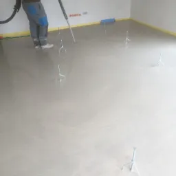 Aplikacja wylewki poziomującej w pomieszczeniu, widoczne metalowe statywy poziomujące i wąż podający masę, zabezpieczone ściany taśmą.