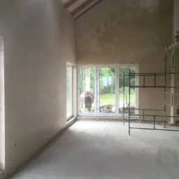 Wnętrze domu w trakcie prac wykończeniowych: ściany po nałożeniu tynku, surowa podłoga, widoczne rusztowanie i mieszalnik do tynku za oknem.