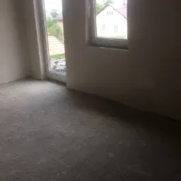 Surowa posadzka w trakcie wykonywania w jasnym, surowym wnętrzu z dwoma oknami, widok na dom i zieleń na zewnątrz.