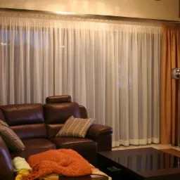 Salon z brązową skórzaną kanapą, stolikiem kawowym i oknem zasłoniętym brązowymi i białymi, delikatnymi firanami.