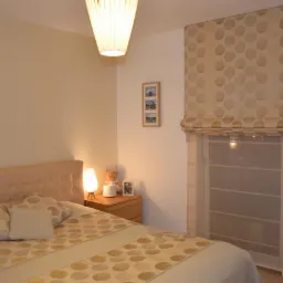 Beżowa roleta rzymska w kropki, zamontowana w oknie sypialni z beżowym łóżkiem i lampką nocną