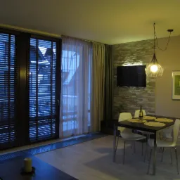 Jadalnia z ciemnymi drewnianymi żaluzjami poziomymi na oknach balkonowych, jasnymi zasłonami i kamienną ścianą z telewizorem.