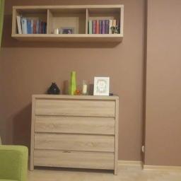 Piotr Kijewski - Beżowa ściana po malowaniu z drewnianą komodą, półką na książki i dekoracjami, widoczny fragment zasłony i sofy.