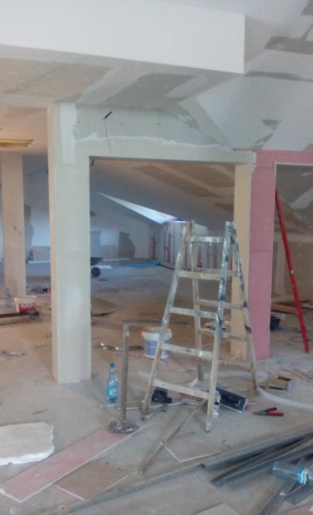 Wnętrze w trakcie prac wykończeniowych: widoczne szpachlowane ściany i sufit, drewniana drabina malarska, butelka wody i narzędzia na podłodze, filar w surowym stanie.