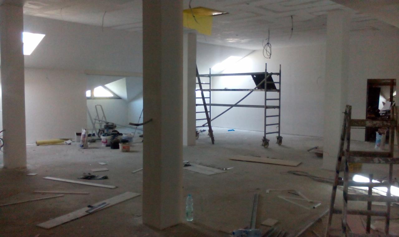 Wnętrze w trakcie prac wykończeniowych: widoczne rusztowania, kolumny, resztki materiałów budowlanych na podłodze, okna dachowe, oraz gładkie, białe ściany przygotowane do dalszych prac.