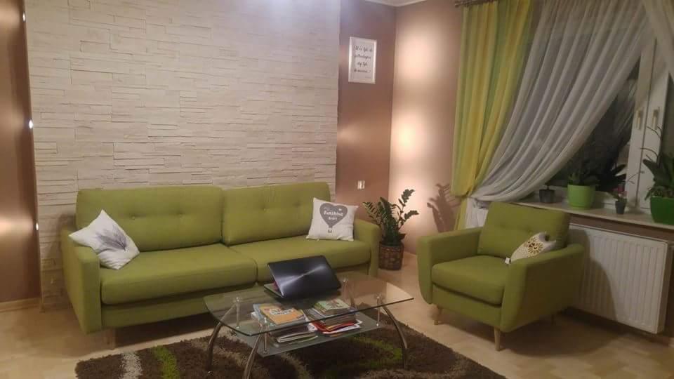 Jasne wnętrze salonu z zieloną sofą i fotelem, ozdobną ścianą z imitacji kamienia, szklanym stolikiem kawowym z laptopem i magazynami, oraz roślinami doniczkowymi przy oknie z żółto-białymi zasłonami.