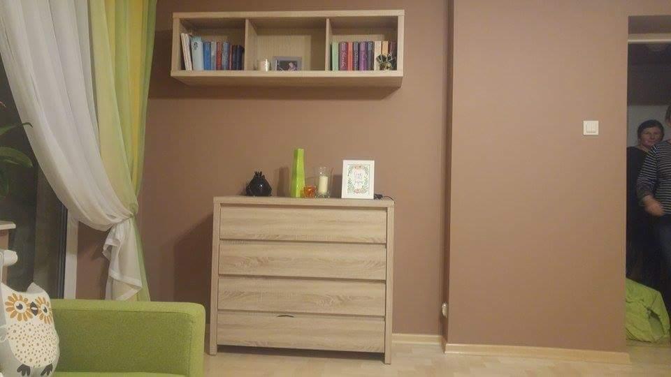 Beżowa ściana po malowaniu z drewnianą komodą, półką na książki i dekoracjami, widoczny fragment zasłony i sofy.