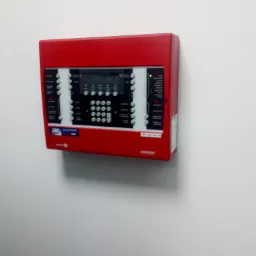 Czerwona centrala alarmowa z wyświetlaczem LCD i przyciskami, zamontowana na białej ścianie, widoczny adres IP.