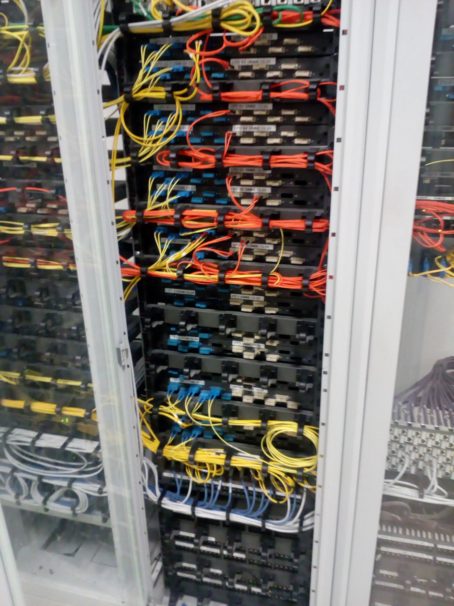 Serwerownia z uporządkowanymi kablami w kolorach żółtym, pomarańczowym, niebieskim i białym, zamocowanymi do czarnych paneli w szafie rack.