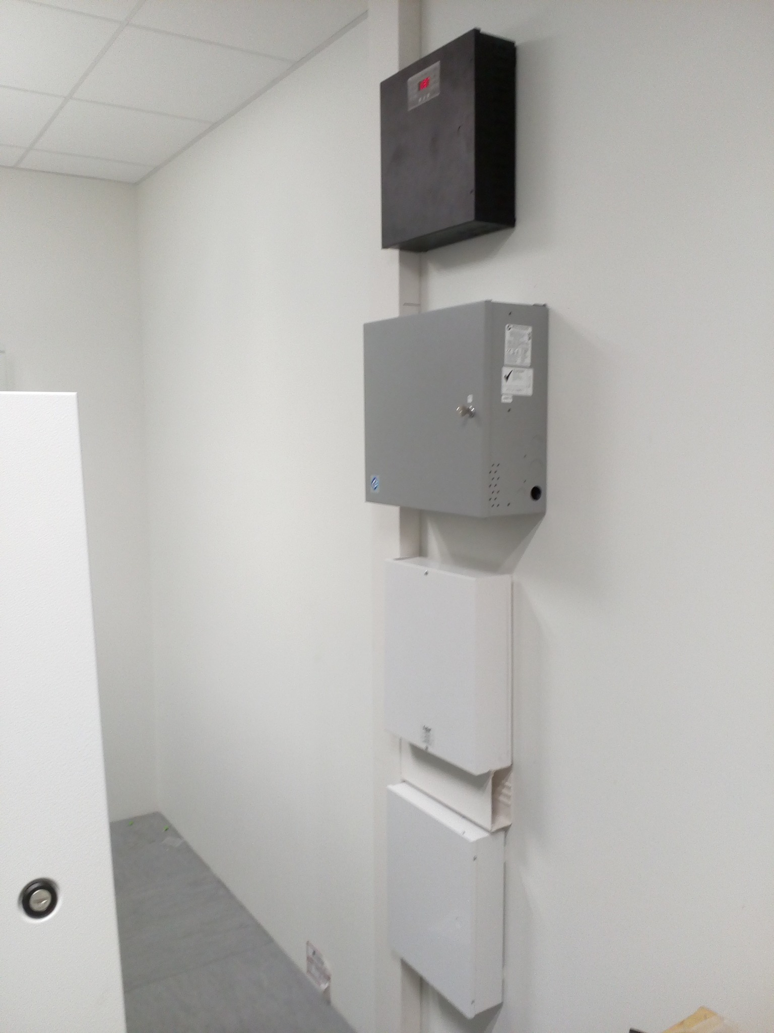 Cztery skrzynki elektryczne, w tym jedna z wyświetlaczem, zamontowane pionowo na białej ścianie wewnątrz pomieszczenia. Widoczna część podłogi i fragment białych drzwi po lewej stronie.