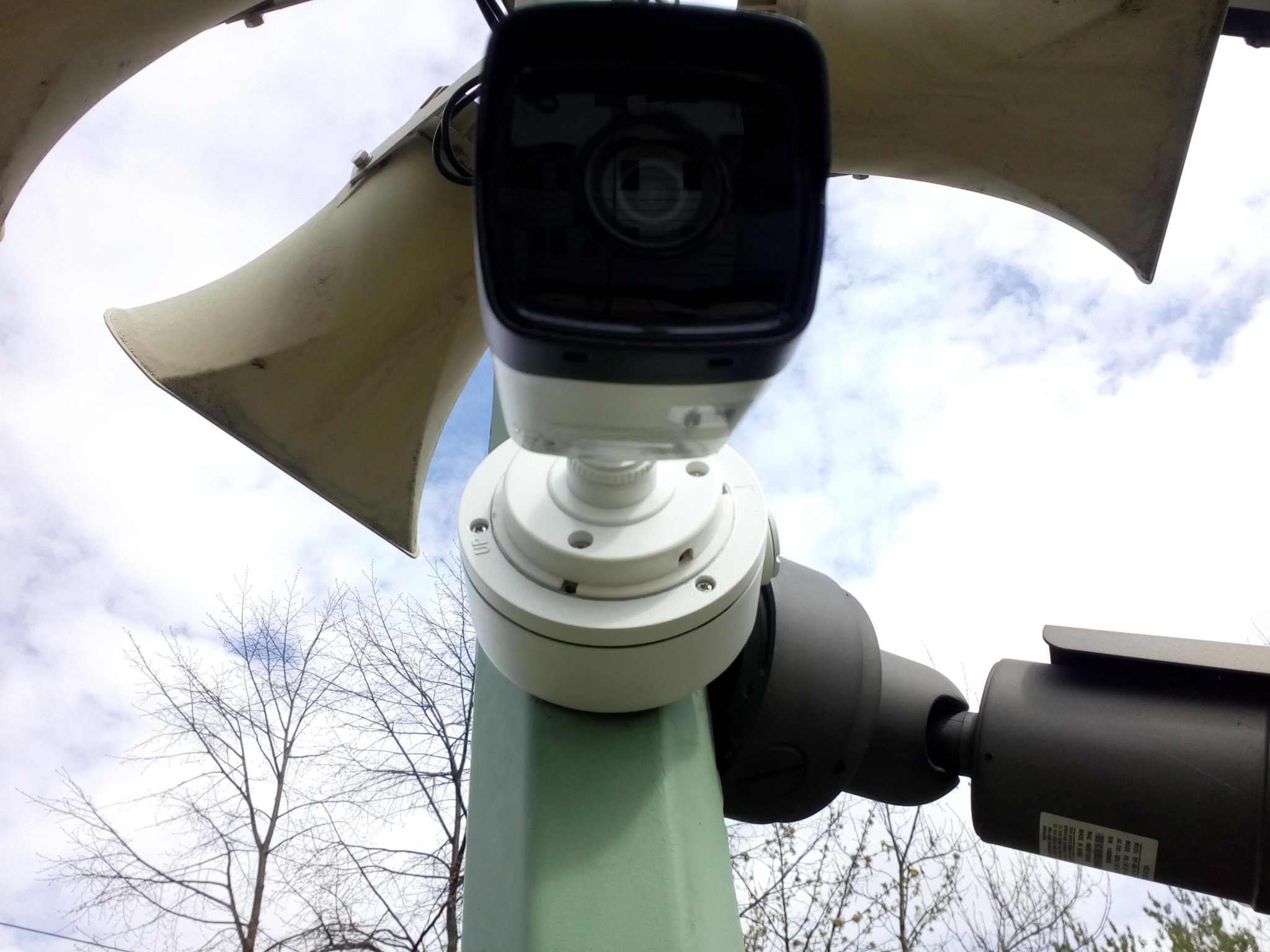 Kamera monitoringu i głośniki tubowe zamontowane na słupie, widok z dołu na tle błękitnego nieba z chmurami i gałęzi drzew.