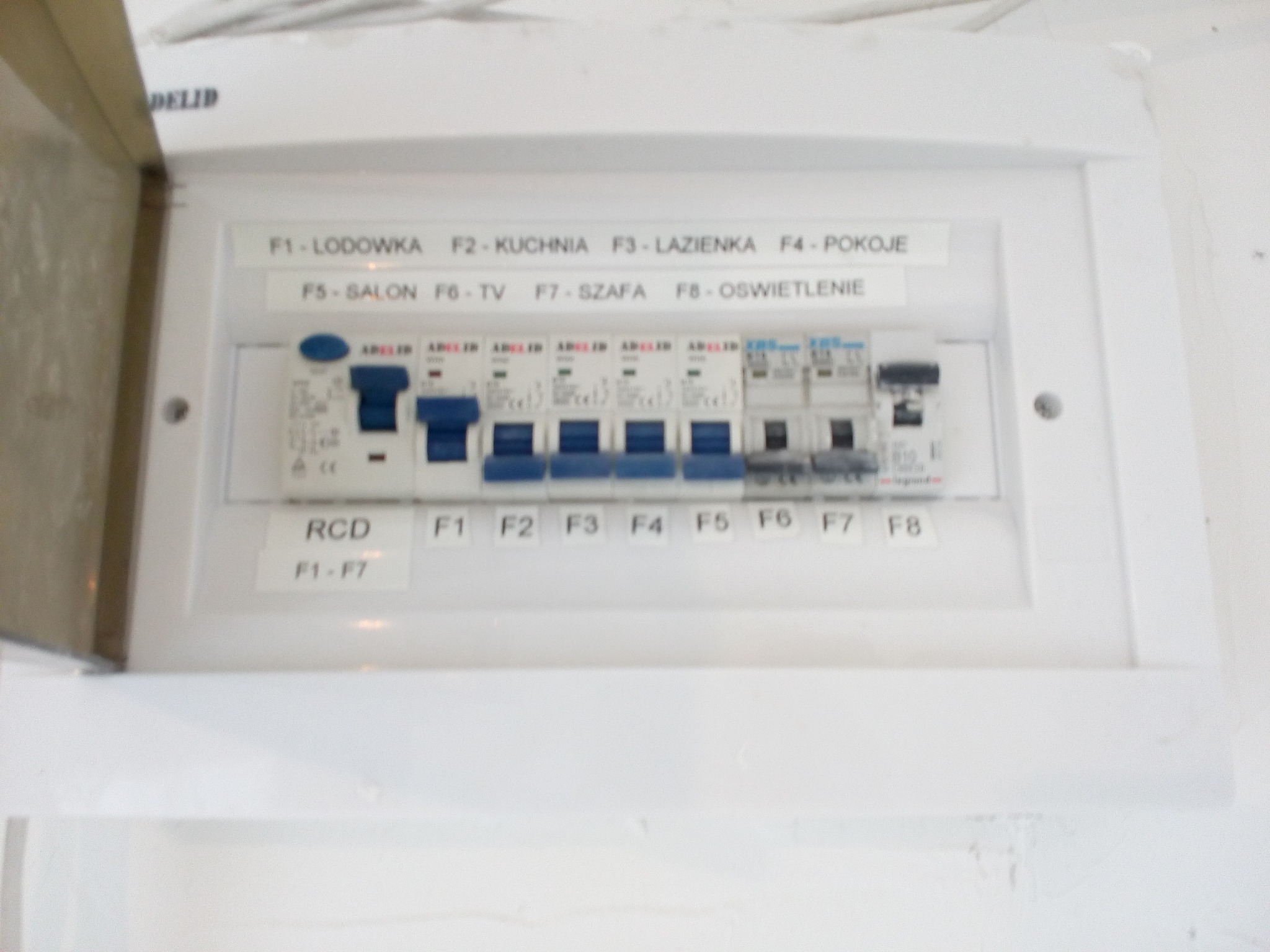 Otwarta skrzynka bezpiecznikowa z opisem obwodów: lodówka, kuchnia, łazienka, pokoje, salon, TV, szafa, oświetlenie.