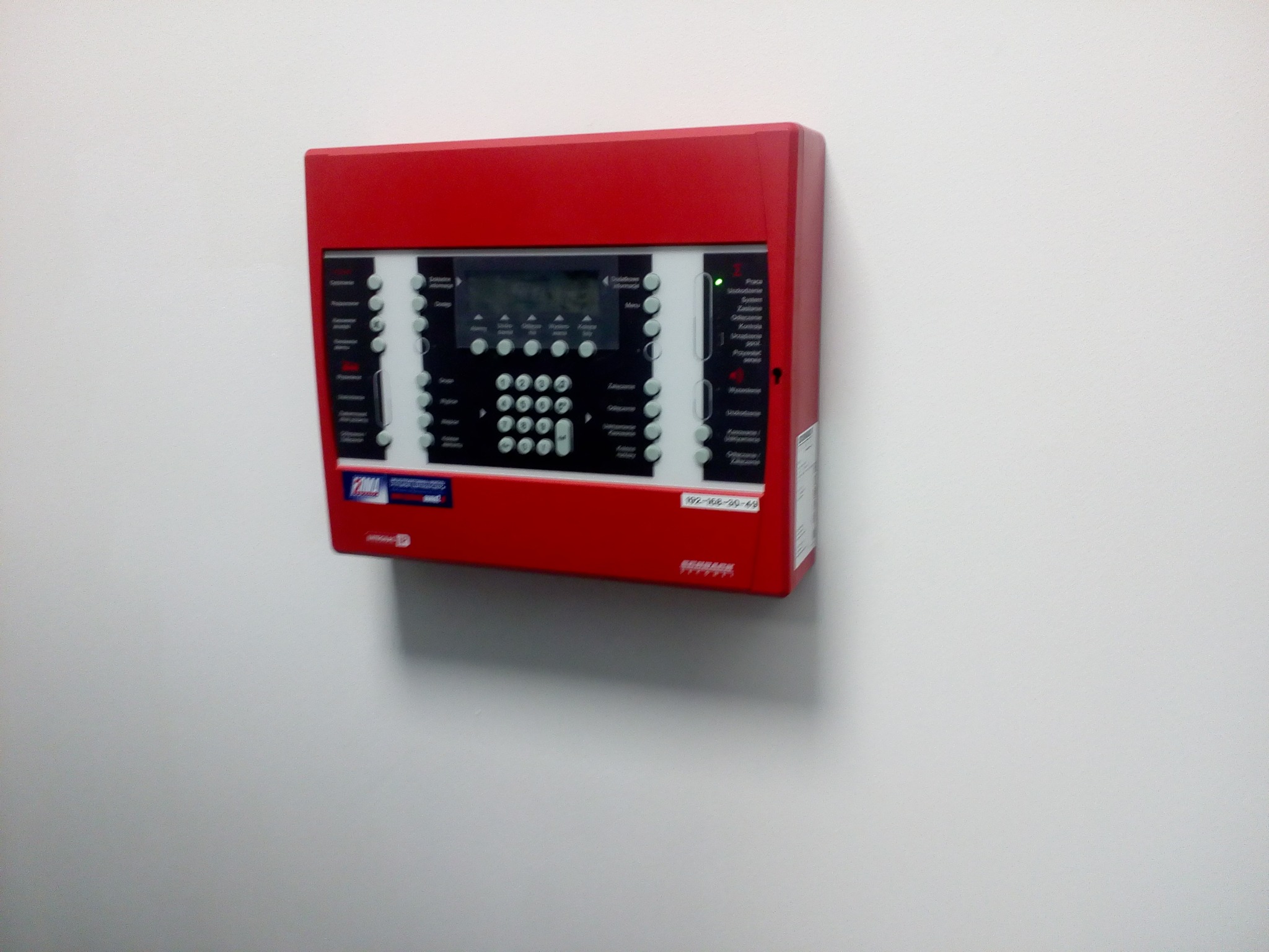 Czerwona centrala alarmowa z wyświetlaczem LCD i przyciskami, zamontowana na białej ścianie, widoczny adres IP.