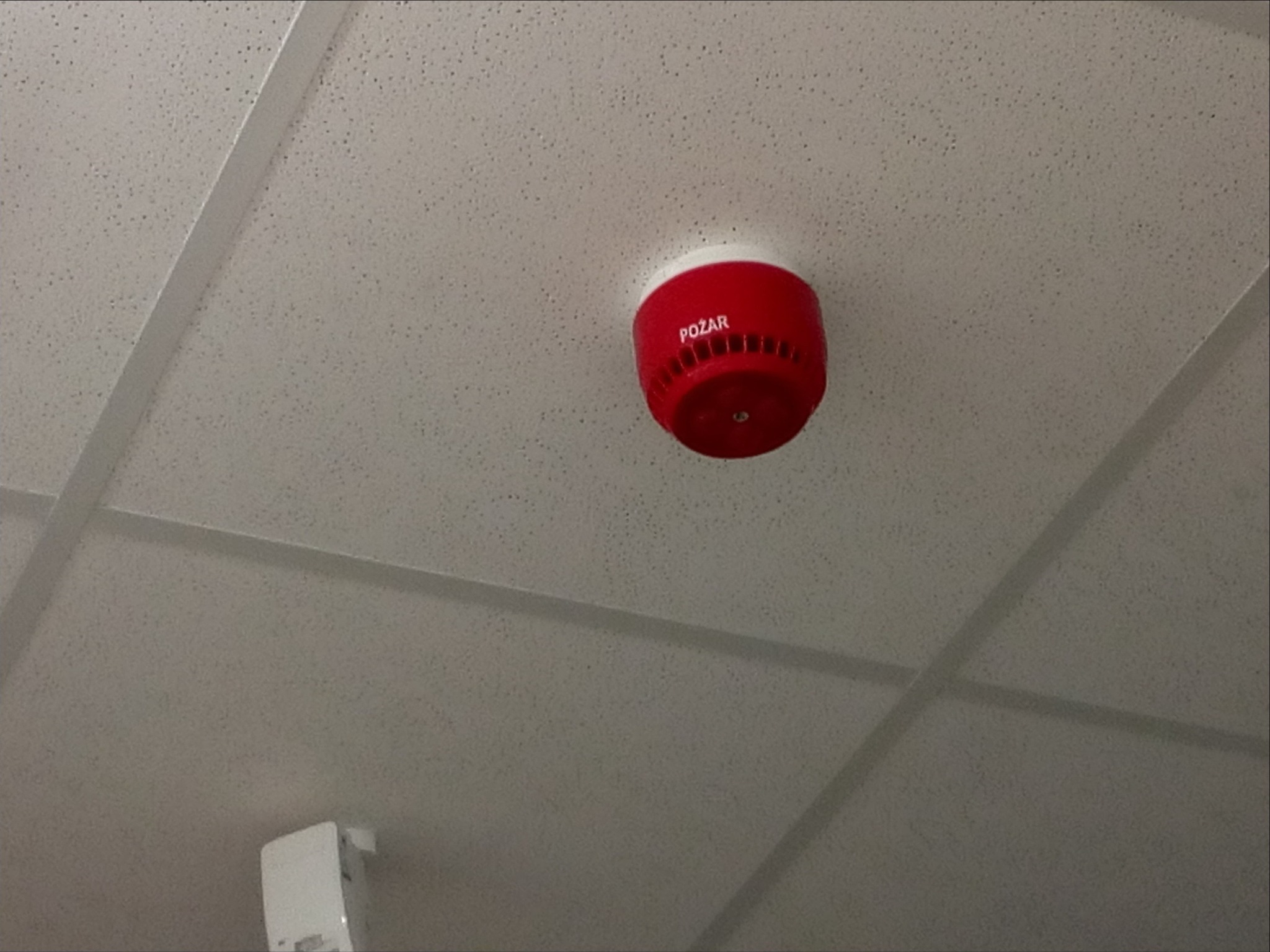 Czerwony czujnik dymu z napisem 'POŻAR' zamontowany na białym suficie podwieszanym z widocznymi elementami konstrukcyjnymi.