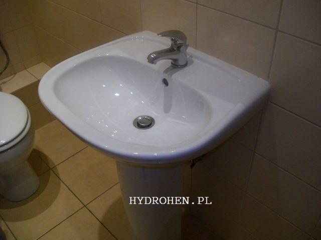 Umywalka ceramiczna na postumencie z chromowaną baterią, widok z boku, w tle beżowe płytki i toaleta.