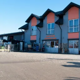 Fasada budynku Centrum Budowlanego z widocznymi materiałami budowlanymi na zewnątrz, w tym ogrodzenia, rury i kostka brukowa, na pierwszym planie ułożona kostka brukowa.