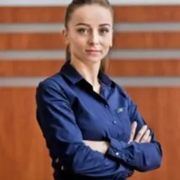 Portret kobiety w niebieskiej koszuli z założonymi rękami na tle drewnianej ściany z poziomymi paskami.