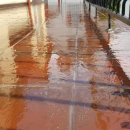 Mokry, wykafelkowany taras z metalową balustradą, po deszczu, z widocznymi odbiciami elementów budynku.