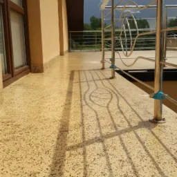 Wykończony balkon z beżową, nakrapianą posadzką i chromowaną balustradą z dekoracyjnym wzorem, częściowo jeszcze zabezpieczoną folią. W tle widoczne zachmurzone niebo.