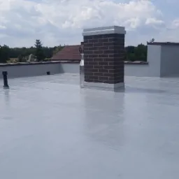 Świeżo pomalowany, jasnoszary płaski dach z ceglanym kominem i odpowietrznikami, widok z góry w słoneczny dzień.