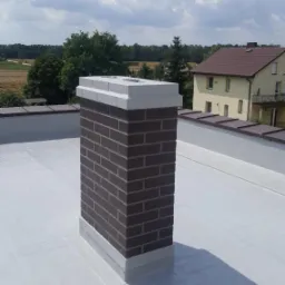 Biały, płaski dach z ceglanym kominem wykończonym szarymi elementami, w tle budynek mieszkalny i zielone drzewa pod błękitnym, zachmurzonym niebem.