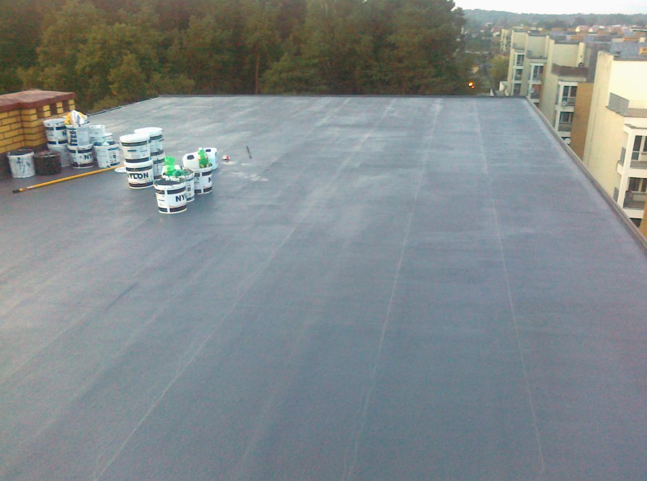 Płaski dach pokryty szarą membraną bitumiczną, widoczne puszki z klejem i narzędzia, w tle zabudowa miejska i zieleń.