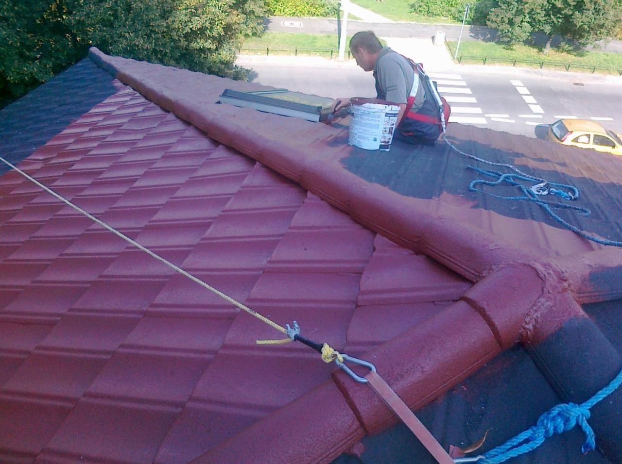 Pracownik w uprzęży maluje dachówkę na dachu, widoczny fragment pomalowanej powierzchni i puszka z farbą, w tle ulica i drzewa.