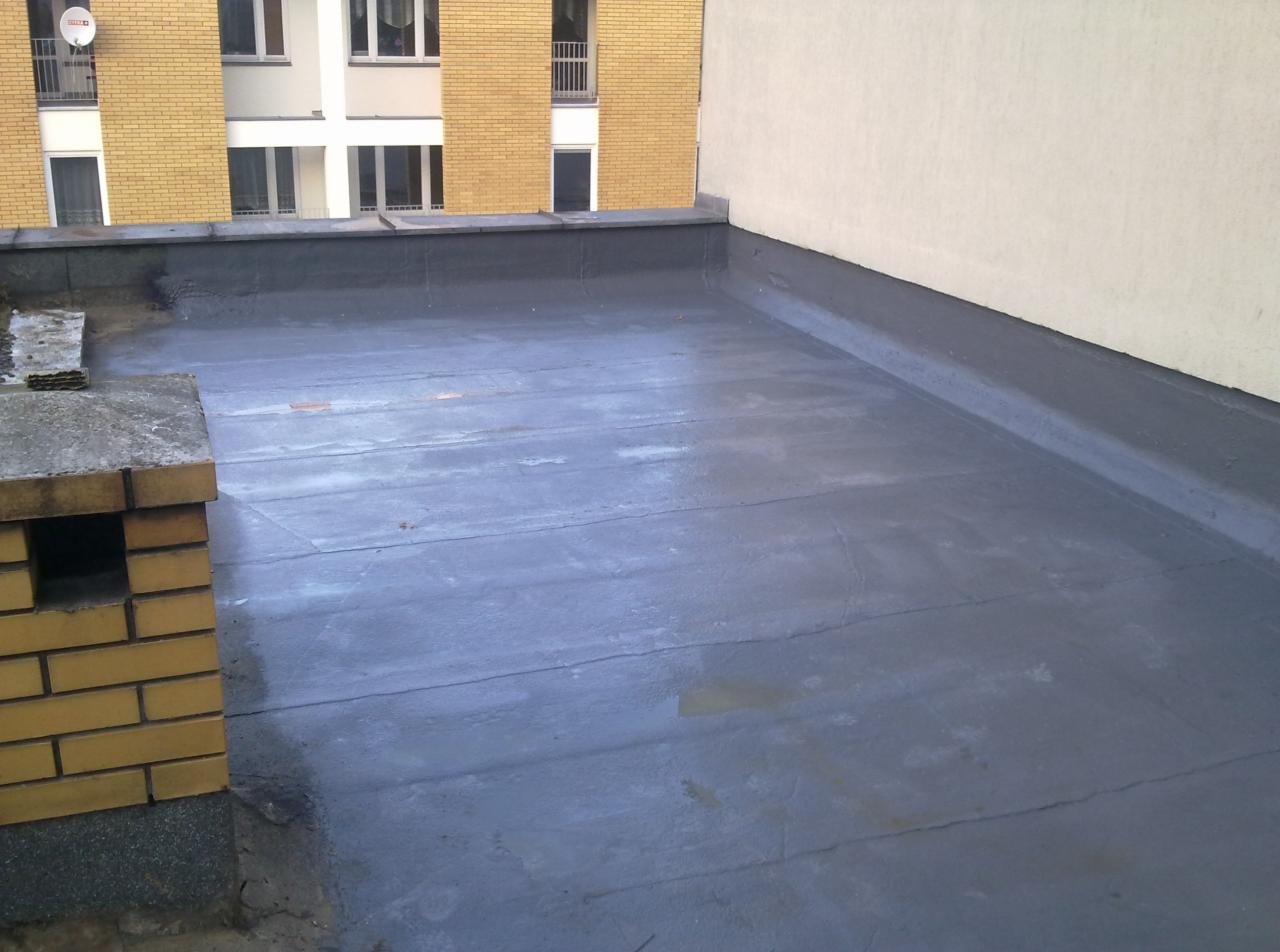 Płaski dach pokryty szarą papą bitumiczną, widoczny komin z cegły klinkierowej i fragment budynku w tle.