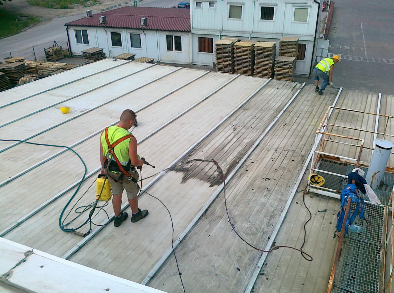 Dwóch pracowników w kamizelkach odblaskowych czyści dach z blachy trapezowej za pomocą myjki ciśnieniowej, widoczne zabrudzenia i resztki brudu na powierzchni dachu.